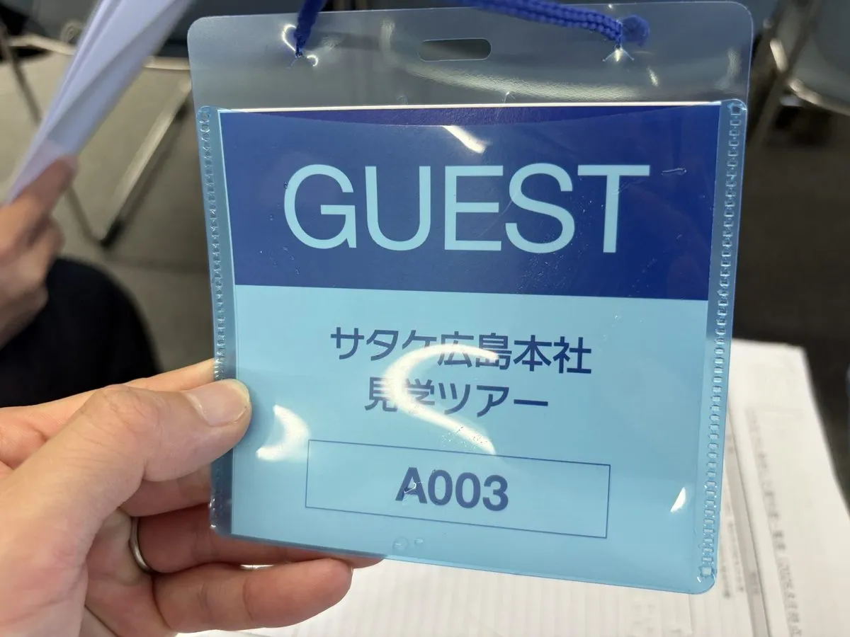 「GUEST サタケ広島本社見学ツアー A003」と書かれた青いゲスト証を手に持っている